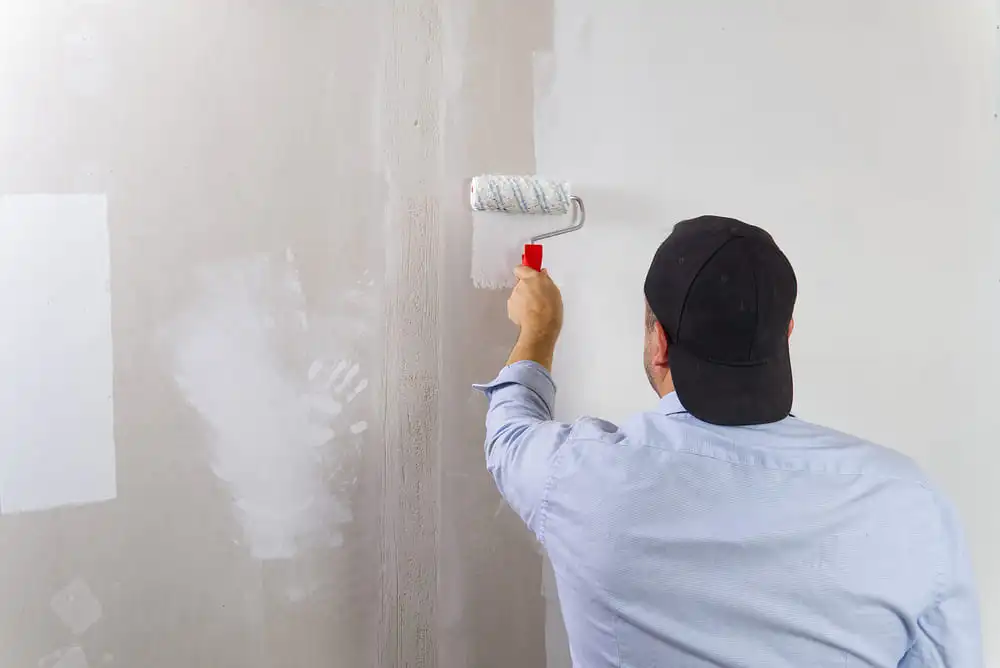 Painter applying white paint to repaired drywall wall indoors
